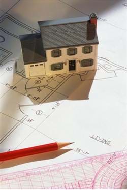 A model of a house and the accompanying blueprint. Shown are a miniature, 2-story house, sitting on a blueprint, and next to a pencil and architect's ruler.