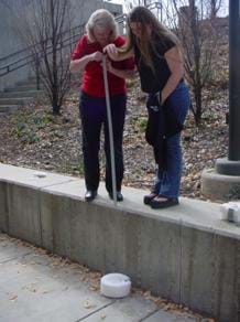A photograph shows an outside egg drop testing station. Two women stand on a bench. One holds a yardstick while the other is about to release an egg from about six feet above a prototype foam landing pad.
