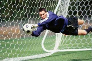 Action photograph of a soccer goalie stopping a shot on goal. The goalie appears to be flying through the air to stop the ball.