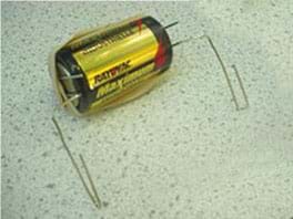 A photo shows a battery with a looped paperclip on each end. A rubber band stretches lengthwise across the battery to secure the two paperclips.