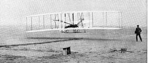 Black and white photograph of a simple bi-wing aircraft with one man lying face down, head forward in the center of the open plane and a second running nearby.