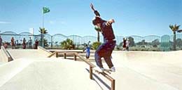 A photograph showing a skateboarder doing a rail slide at a public skate park in San Diego. With both hands up in the air for balance, he stands with both feet on his skateboard, which is on the top of a horizontal hand rail.
