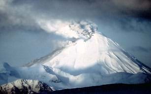 Photo shows white and gray dirt and ash being ejected from the peak of a tall, snow-covered mountain.