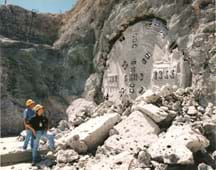 Photo shows two men looking at metal prongs coming through the side of a mountain with a crumbling rock pile in front of them.
