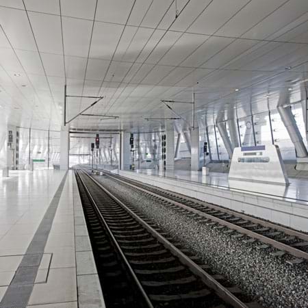 A photo shows the interior tunnel of a modern railway station with two sets of train tracks, ceiling, windows and waiting platforms. 