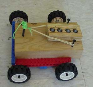 A photograph shows a block of wood placed on a LEGO-type platform onto which four small rubber wheels are attached. A second, smaller wooden block with weights (fishing sinkers) resting inside four holes in the block rests on the bigger wooden block, and will be launched off the back of the vehicle via a rubber band.