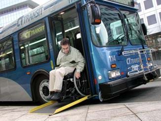 A photograph of man in a wheelchair who is exiting a public city bus via a ramp. One end of the ramp is placed on the bus steps, while the opposite end is placed on the sidewalk.