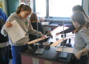 A photograph of students at lab table preparing to measure their cars pulling weight up an inclined plane. Shown are two wood planes attached with a clamp to a stand.