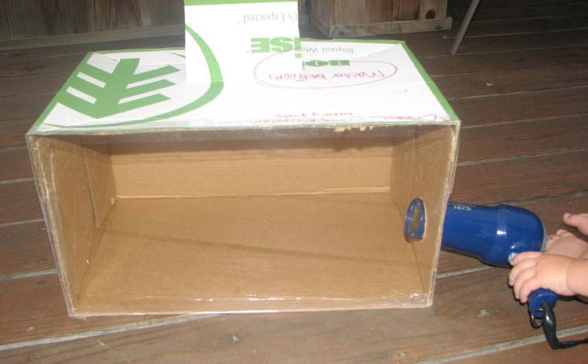 Photo shows a closed cardboard box with one side replaced with clear Plexiglas, and hairdryer nozzle inserted into round hole on one side.