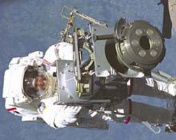 Photo shows a person in a white space suit floating by some equipment, with the surface of the Earth as the background.