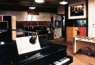 Photo shows a room with a piano, instruments and microphones  in the foreground along with other musical instruments. 