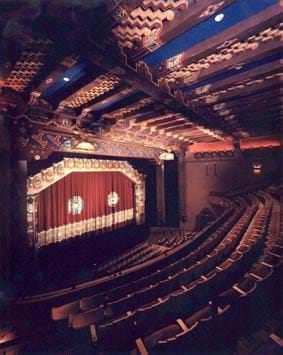 A photograph of the KiMo Theatre in Albuquerque, NM. Shown are rows of curved seats, each row rising above the previous row, a stage with a red curtain and corrugated panels on the ceiling.