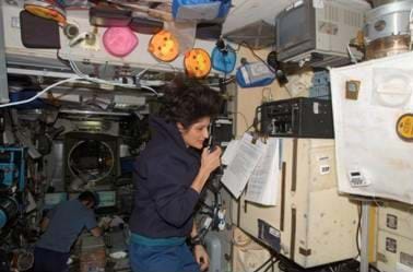 Photo shows a female astronaut talking on a handheld device while floating in a scientific laboratory. Another astronaut in the background is collecting data on a computer.