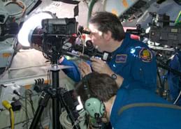 Photo shows two men in blue jump suits operating what looks like a large camera on a tripod facing out a porthole window.