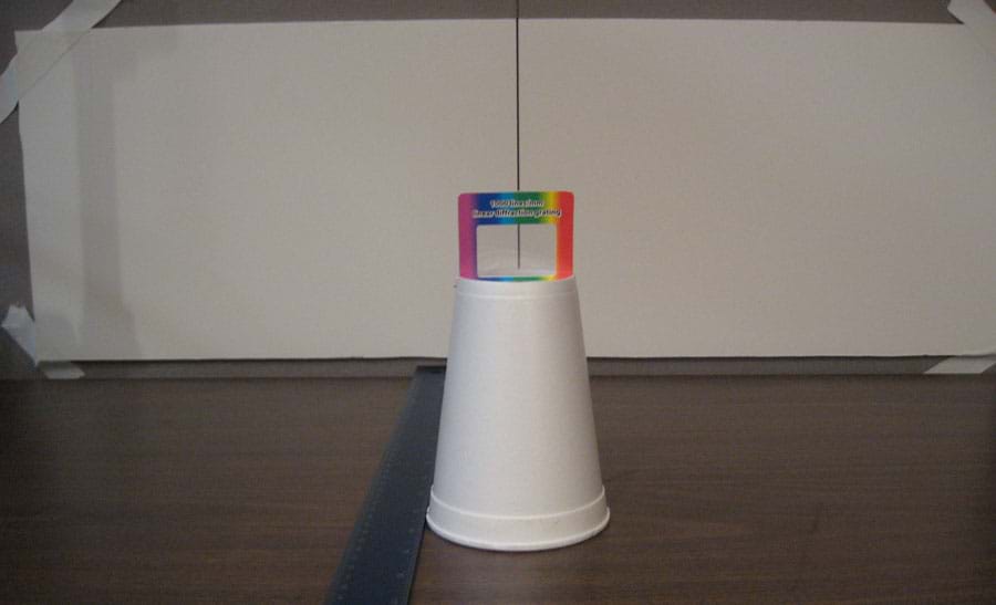 Photo shows an upside-down cup on a tabletop, lined up in front of a black vertical line in the middle of a wide white paper taped to the wall behind, with a ruler positioned on the table nearby.
