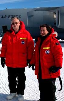 Photo shows two adults wearing bright red parkas as they stand near an Air Force jet parked on snow-packed ground.