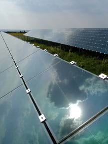 Photo shows an array of solar panels with the sun, sky and clouds reflected in them. 
