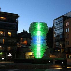 A photograph shows an illuminated green and blue (somewhat) tornado-shaped five-story light sculpture in a courtyard with seven-story buildings nearby. 