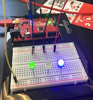 A photograph shows a breadboard connected to a SparkFun RedBoard microcontroller. The breadboard has three circuits in it, each with a resister and LED, and each wired to the red circuit board. The green and blue LEDs are illuminated; the yellow LED is not.