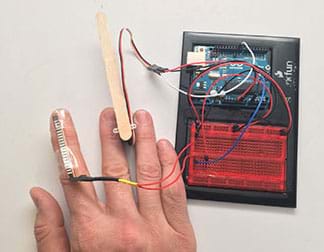 A photograph shows a flex sensor taped to the index finger of hand, which is connected to a breadboard and Arduino microcontroller. A Popsicle stick held between the hand’s middle and ring fingers is also connected to the Arduino.