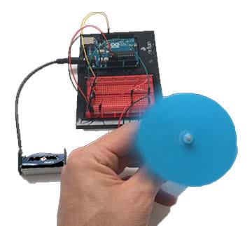 A photograph shows a hand holding a blue spinning paper fan that is attached to a motor controlled by an Arduino microcontroller, breadboard, wires and battery that are seen in the background.