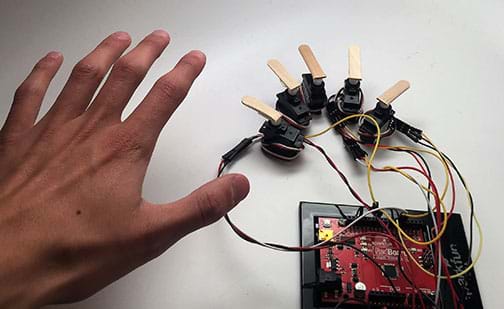 A photograph shows five servos each connected by wire to an Arduino. On each servo is hot-glued a portion of a popsicle stick, making each like a finger, and together, mimicking a human hand. For comparison, an open hand is next to the five servos. 