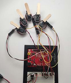 A photograph shows an Arduino and breadboard hooked up to five servo motors, each with glued-on partial Popsicle stick heads, mimicking a five-fingered human hand. Same setup as Figure 3.