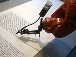 A photograph shows the end of an orange industrial robot arm that is writing text with an ink pen on a roll of paper.