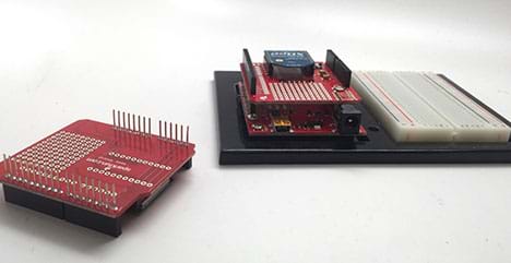 A photograph shows an Arduino microcontroller with an attached breadboard. An Arduino shield is stacked on top of the Arduino board so the pins on the shield are pushed into the pins on the Arduino.