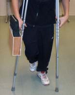 A photo shows a boy using a pair of crutches. Attached to the outer side of one crutch below the hand support is a container—a flat wooden box with the top side open; it contains a notebook.
