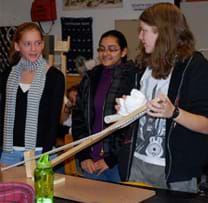 A photograph shows three girls testing the catapult they made from wooden boards, PVC pipes, a hinge and string.