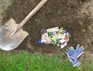 Photo shows view from above of a hole in the ground filled with assorted trash (vegetable peelings, packaging wrappers, tissue, egg shells, tea bag, can lid, plastic lid), and a shovel and gloves nearby.