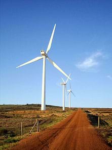A photograph shows three very tall, three-bladed white wind turbines along a rural dirt road in Cape Town, South Africa.