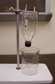 Photo shows the top half of a plastic beverage bottle suspended narrow-neck down above a glass beaker via plastic ties attached to a horizontal rod clamped to a vertical post in a base.