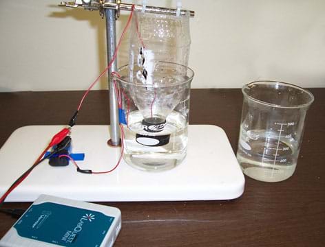 Photo shows a desktop set-up with the top half of a plastic beverage bottle suspended upside down into fluid in a glass beaker with wires running from inside the bottle and beaker to the battery, resister and probes.