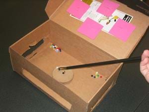 Inside an open box, you can a device holding a bead near a small hook.