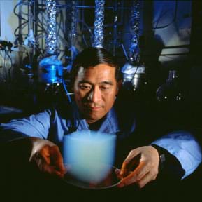 A man (Peter Tsou) in a darkened laboratory uses two hands to hold a plate in front of him. On the plate rests a ~6 x 6-inch cube of aerogel that looks like a floating cube-shaped bluish-milky white cloud (blurry edges).