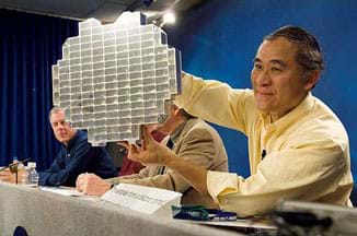 A photograph at a press conference shows a man (Peter Tsou) at a table with three other men, holding up what looks like a shiny, silver round and flat device composed of deep rectangular cells (a stardust sample tray), into which aerogel blocks are placed.
