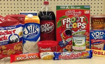 Food items on a grocery shelf: Cracker Jack caramel popcorn in a bag, soy milk in a carton, soda in a plastic bottle, cereal in a box, cereal in a plastic cup, potato chips (Pringles) in a tall cardboard canister with resealable lid, pop tarts in a cardboard box, candy bars in Mylar and paper wrappers, soup in a plastic bowl with a plastic lid.