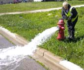 Photo shows a woman using a wrench on a fire hydrant that is spewing water.