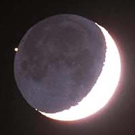 A photograph shows a gray round Moon in a black sky with one arc-ed edge of the moon (a crescent shape) glowing white and bright.