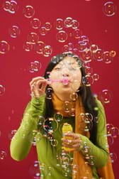 A girl blows through a bubble wand, creating a mass of floating soap bubbles.