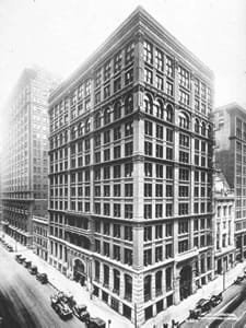 A black and white photograph shows a 14-story bilding on the corder of a city block.
