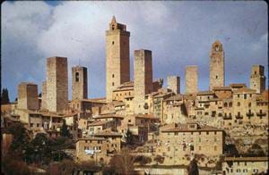 Photo of a town of stone buildings with tile roofs, including many tall towers that rise above the rest of the buildings.