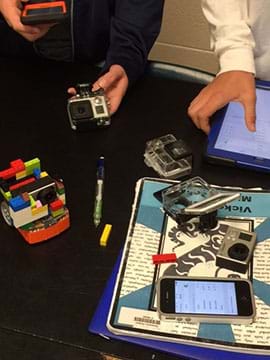 A photograph shows two students setting up a small (Edison) robot and handling a computer tablet, smartphone and digital camera. LEGO bricks on the robot are positioned to hold the camera.