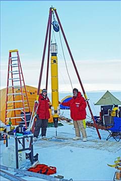 A photograph shows a snowy landscape in Antarctica with two people in red parkas standing on snow-covered ice near a long, cylindrical yellow device hanging from a triangular metal frame above a hole in the ice. They are preparing to deploy the Icefin robot through the hole into the water under the ice.