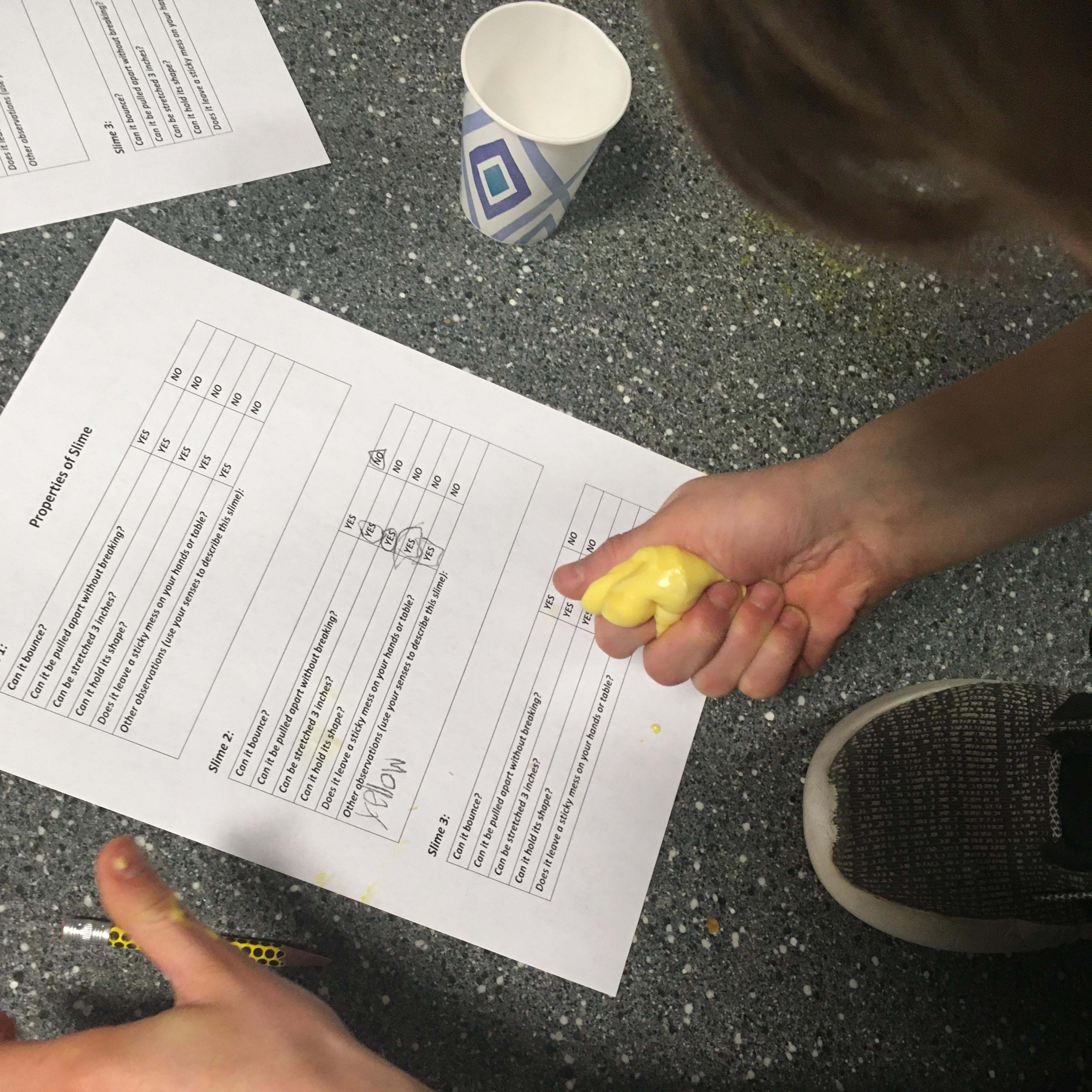 A student holds a yellow slime ball while filling out a worksheet to assess its properties. 