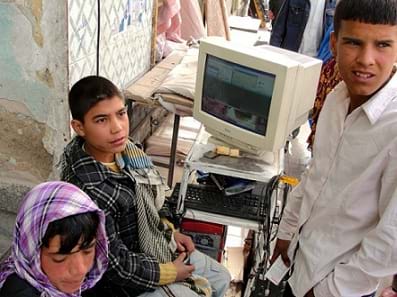 A photograph shows three children near a desktop computer with an older-style box-shaped monitor at an outdoor marketplace in Kabul, Afghanistan.