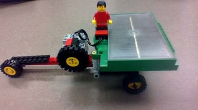 A photograph shows a small-size solar-powered wheeled vehicle made from an eLAB LEGO Renewable Energy Set. It has a long nose with a small wheel at the end and multiple wheels at the back, on which the solar panel is positioned. A LEGO person rides in the middle of the vehicle. 
