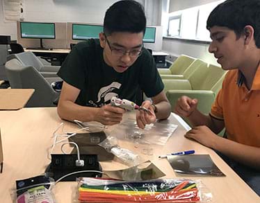 A photograph shows two teens at a classroom table constructing their prototype sunglasses. One student uses a hot glue gun to affix the polarizing filter while the other is assisting. 